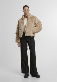 Beežist faux fur puffer jakk, millel on kõrge krae, tõmblukuga sulgemine ja tikanditunne, koos mustade laiade pükstega ja tursketega mustade kingadega.