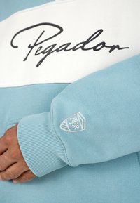 Svetlomodrá mikina s bielym horizontálnym pásom, na ktorom je čiernym písmom logo "Pegador". Rúbkované manžety a malá vyšitá nášivka na rukáve.