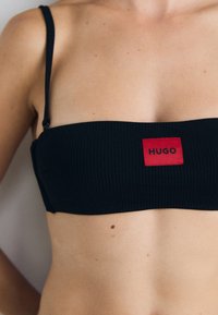 Svart ribbet bandeau-top med tynne stropper, med et rødt rektangulært merke merket "HUGO" i svart tekst i midten.