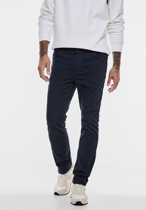 Navy blue slim-fit chinos made of soft cotton, featuring five pockets and a zipper fly, paired with light-colored sneakers.