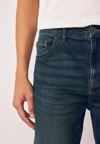 Tumed sinine denimist teksapüks, millel on nähtavad õmblused, eesmine tasku ja tekstureeritud pind, eksponeeritud käsi reiele toetumas.