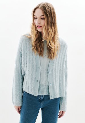 Young woman with long wavy hair wearing a light blue cable-knit cardigan over a matching sweater and blue jeans, looking down.