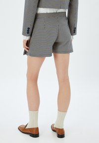 Pantalones cortos a medida con un patrón de cuadros, confeccionados en una mezcla de telas, que presentan una cintura alta, bolsillos laterales y un acabado limpio. Combinados con zapatos marrones y calcetines crema.