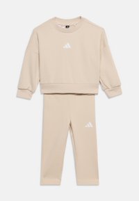 Σετ μπλούζας cropped με μακριά μανίκια σε μπεζ χρώμα και αντίστοιχες κολάν, με λευκό λογότυπο Adidas στο στήθος και στο πάνω μέρος του ποδιού.