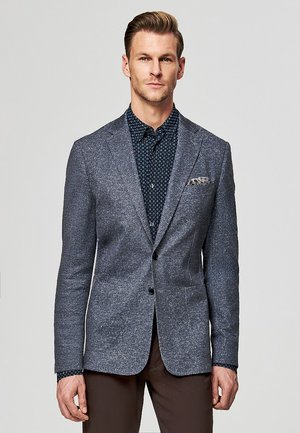 Mężczyzna w niebieskiej, fakturowanej marynarce na ciemnej, wzorzystej koszuli, brązowe spodnie, stojący na jasnym, jednolitym tle.