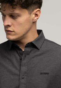 Młody mężczyzna z krótko przyciętymi, kręconymi włosami, w ciemnoszarej koszuli z guzikami z logo "Lee Cooper" na piersi, patrzący lekko w dół w lewo.