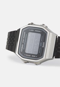 Montre numérique Casio avec boîtier rectangulaire en argent, bracelet en silicone noir, affichage LED affichant l'heure, la date et le nombre de pas, résistant à l'eau.