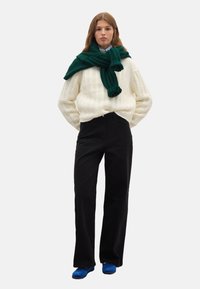 Cremefarbener Strickpullover, grüner Schal, schwarze weit geschnittene Hose und blaue Schuhe. Weiche Texturen und lockere Passform im gesamten Outfit.