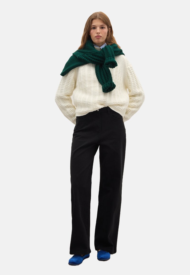 Cremefarbener Strickpullover, grüner Schal, schwarze weit geschnittene Hose und blaue Schuhe. Weiche Texturen und lockere Passform im gesamten Outfit.