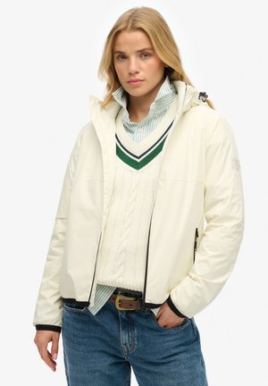 Witte geïsoleerde jas met zwarte accenten, ritssluiting aan de voorkant en een capuchon. Gelaagd met een crèmekleurige kabelbrei trui en een lichtblauwe gestreepte blouse eronder.