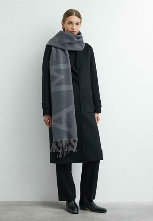 Woman wearing a long black coat, black pants, black ankle boots, and a large gray scarf with fringe standing against a plain white wall.
