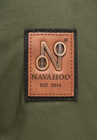 Brun läderetikett med präglad logotyp 'N' och 'NAVAHOO EST 2014', omgiven av svart tyg, sytt på grön textilbakgrund.
