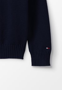 Manica di un maglione lavorato a maglia blu navy con polsino a costine. Caratterizzato da un piccolo logo tricolore (rosso, bianco, blu) nella zona del polso.
