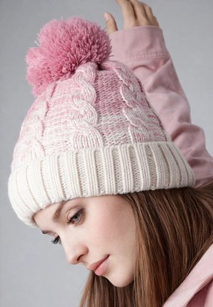 Young woman wearing a pink and white knitted hat with pom-pom, looking down, with one hand raised behind her head.