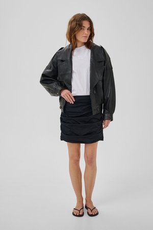 Vrouw die een zwart leren jasje draagt, een wit T-shirt, geplooide zwarte rok en zwarte teenslippers staat tegen een effen grijze achtergrond.