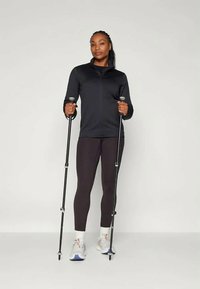 Μαύρο φανελάκι με φερμουάρ, μαύρα κολάν και γκρι αθλητικά παπούτσια με πορτοκαλί λεπτομέρειες. Κρατά δύο ραβδιά πεζοπορίας, στέκεται μπροστά σε απλό φόντο.