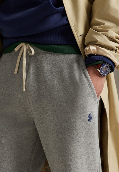 Dettagli di un outfit casual con pantaloni della tuta grigi con coulisse, logo blu, manica di cappotto beige e orologio da polso sulla mano sinistra in tasca.