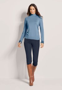 Frau trägt ein blaues langärmeliges Oberteil mit Stehkragen und gemusterten Bündchen, schwarze schmale Hose und braune kniehohe Stiefel, steht vor einem schlichten Hintergrund.