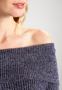 Femme portant un pull en maille bleu marine à épaules dénudées, montrant son épaule nue et une partie de son visage souriant.