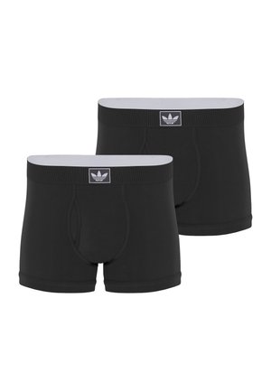 Moški brez majice stoji bos, nosi črne Adidas boxer hlačke, prilagaja pas, gleda naprej na belem ozadju.