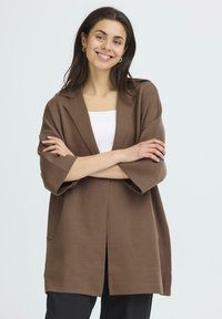 Ruda didelė kardiganas su apykakle, 3/4 ilgio rankovėmis, atviru priekiu ir minkšta tekstūra. Dėvimas ant balto marškinėlio, derinamas su juodomis kelnėmis.