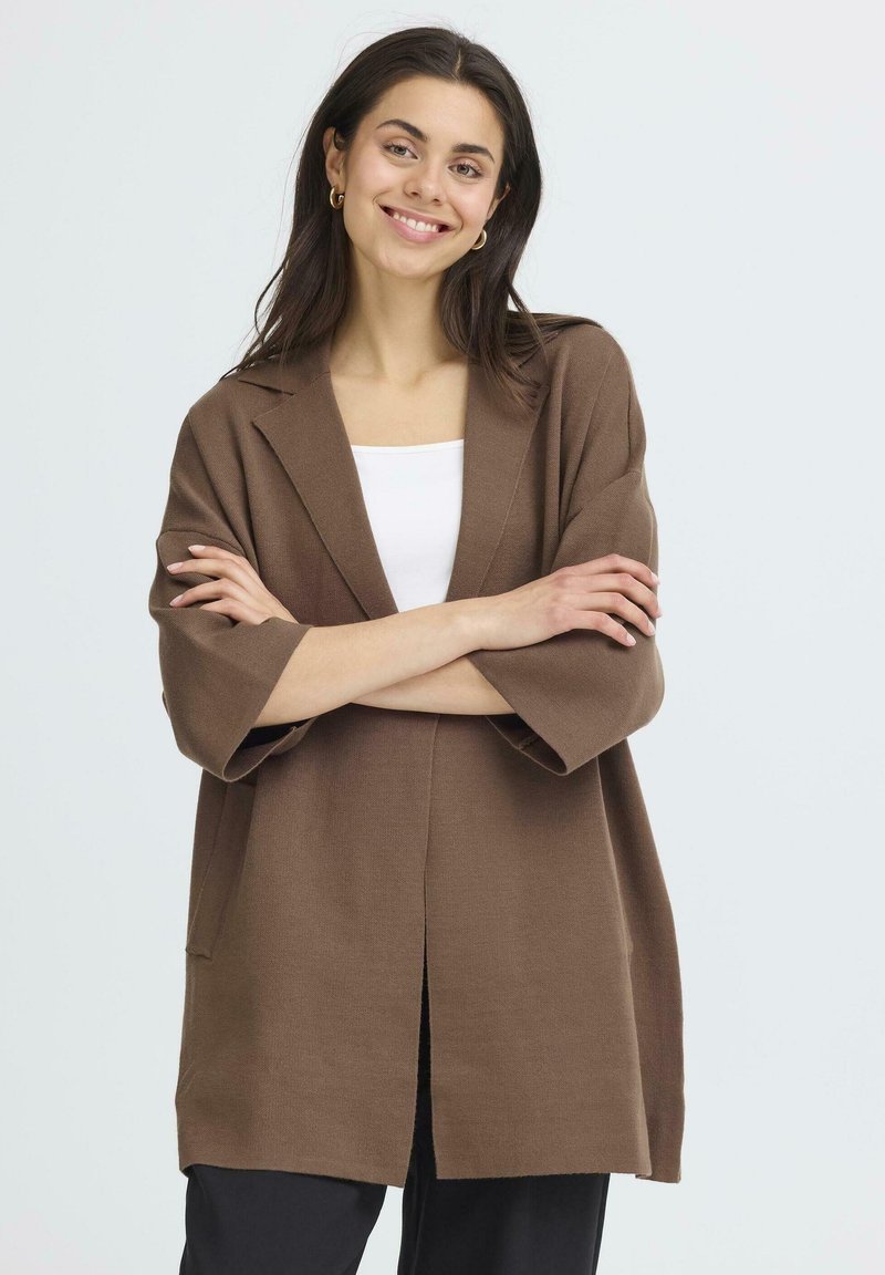 Ruda didelė kardiganas su apykakle, 3/4 ilgio rankovėmis, atviru priekiu ir minkšta tekstūra. Dėvimas ant balto marškinėlio, derinamas su juodomis kelnėmis.