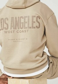 Sudadera beige con gran texto "LOS ÁNGELES COSTA OESTE". Cuenta con capucha con cordones y dobladillo acanalado, con coordenadas y "CALIFORNIA" debajo.
