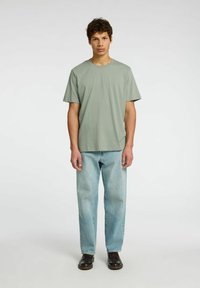Camiseta verde claro de manga corta combinada con jeans rectos azul claro y zapatos marrones, de pie sobre un fondo neutro.