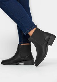 Musta nahkasaapas nilkkapituisilla joustavilla sivupaneeleilla, matalalla lohkareella ja sileällä pinnalla, yhdistettynä tummansinisiin istuviin farkkuihin.