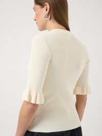 Femme portant un pull crème côtelé à manches courtes avec des poignets à volants et des boucles d'oreilles argentées en forme d'anneau, vue de dos et de profil.