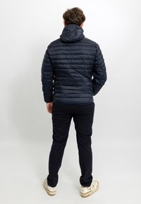 Giacca puffer blu scuro con design trapuntato, cappuccio incorporato e tessuto elasticizzato, abbinata a pantaloni scuri e scarpe da ginnastica di colore chiaro.