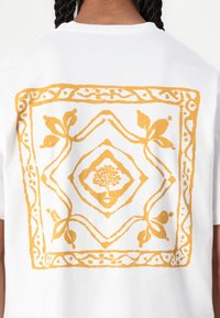 Camiseta de algodón blanca con un gran diseño gráfico amarillo en la parte posterior, con un motivo floral y bordes geométricos como detalles destacados.