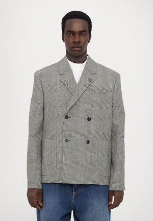 Double-breasted blazer made of houndstooth fabric, featuring black buttons, peaked lapels, and a relaxed fit. Worn over a white shirt.