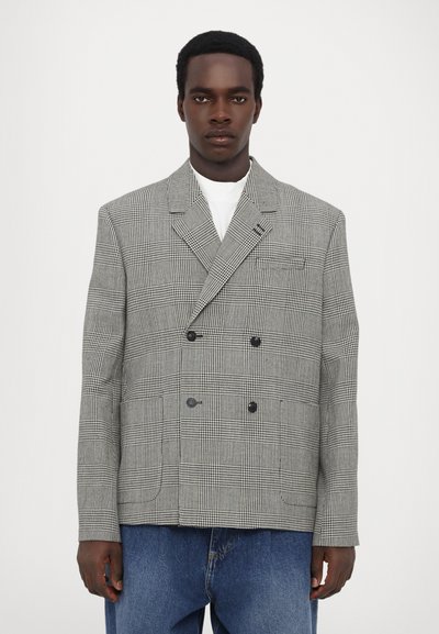Dvojřadý blazer vyrobený z tkaniny s houndstooth vzorem, s černými knoflíky, špičatými klopami a volným střihem. Nosič má na sobě bílou košili.