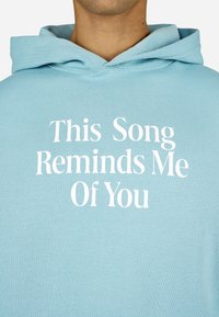 Felpa blu chiaro realizzata in morbido tessuto con testo bianco: "Questa canzone mi ricorda di te," situato in modo evidente nella parte anteriore.