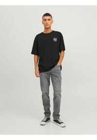 Άνδρας με κοντά μαλλιά, φοράει μαύρο γραφικό μπλουζάκι, γκρι τζιν και μαύρα αθλητικά παπούτσια με λευκές κορδόνες, στέκεται μπροστά σε έναν απλό ανοιχτό φόντο.