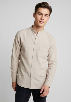 Jonge man die een beige overhemd met mandarijnkraag en lange mouwen draagt en een zwarte broek, staand tegen een effen lichte achtergrond.