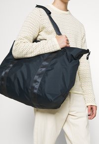 Stor svart nylonväska med dubbla svarta remmar, en mjuk struktur och dragkedjestängning. Har en slät textur utan synliga mönster.