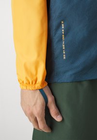 Chaqueta de manga larga con una manga naranja, cuerpo azul con un patrón texturizado, puños elásticos y una etiqueta con coordenadas.