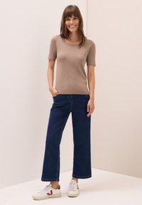 Hnědý top s krátkým rukávem v kombinaci s tmavě modrými širokými džínami a bílými teniskami s červenými detaily; hladká tkanina, uvolněný střih, ležérní design.
