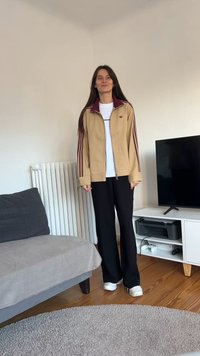 Beige zip-up-jacka med bordeauxfärgad krage och ränder, vit t-shirt, svarta vida byxor och vita sneakers. Rum med en grå soffa och TV.