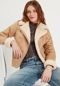 Chaqueta de ante marrón claro con un cuello y puños de piel sintética crema, cierre de cremallera y un corte relajado, combinada con una blusa de estampado floral y jeans azules.