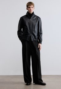 Fekete bőrdzseki magas gallérral, cipzáras záródással és bordázott mandzsettákkal. Lazán szabott fekete nadrággal és fekete cipővel párosítva. Simább textúra.