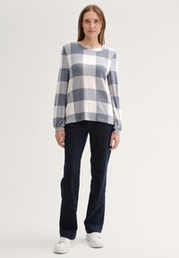 Lichtgrijze en marineblauwe geblokte trui met lange mouwen, gecombineerd met donkerblauwe jeans en witte schoenen, staande tegen een neutrale achtergrond.