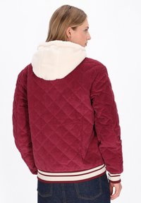 Czerwona kurtka sztruksowa z diamentowym pikowaniem, kremowym kapturem wyścielonym sztucznym futrem oraz ściągaczem z czerwono-białymi paskami.