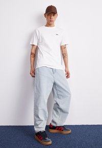 Vit bomullst-shirt med "OBEY"-logotyp, ljusblå, baggy jeans i denim och svarta sneakers med röda ränder och sula i gummi. Brun keps på huvudet.