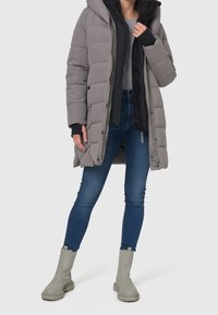 Grå pufferjakke med kviltet design og hætte, lagt over en sort jakke. Båret med blå skinny jeans og lysegrå ankelstøvler.