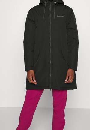 Personne portant un manteau à capuche zippé noir avec des poches latérales et un pantalon magenta, se tenant devant un fond clair et uni.