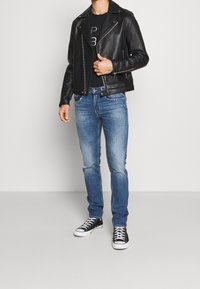 Hombre vestido con una chaqueta de cuero negra, camiseta gráfica negra, jeans azules y zapatillas altas negras de pie contra una pared blanca lisa.