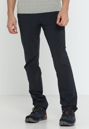 Homme portant un pantalon d'extérieur noir et des chaussures de randonnée noires avec des détails orange, debout sur un sol blanc devant un fond blanc.